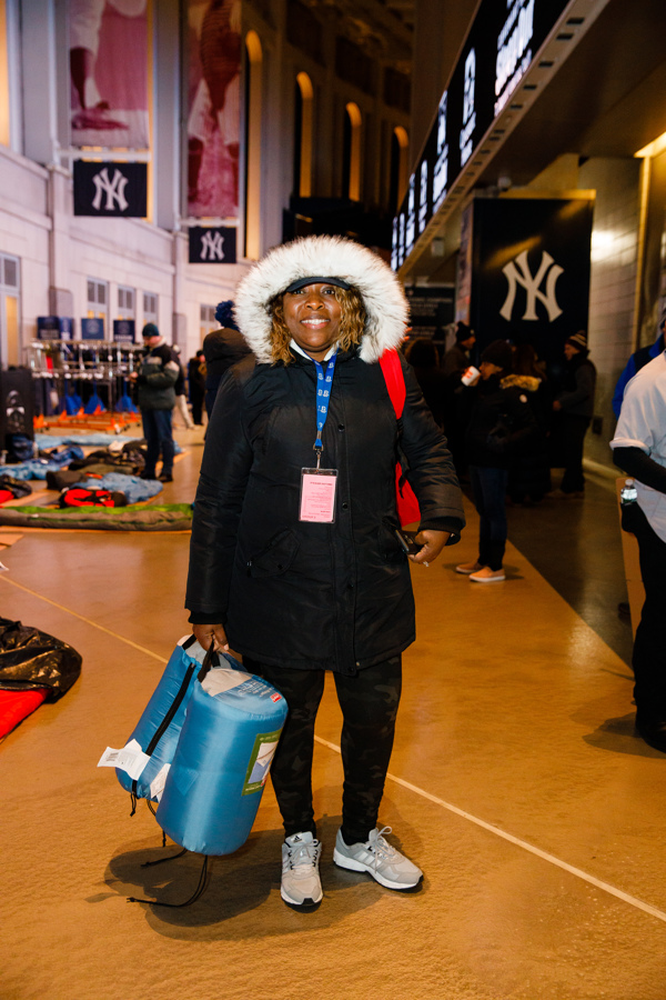 Tracy Jones-Walker in a hooded parka gets ready to Sleep Out in a corridor at Yankee Stadium. Characteristic banners with white N-Y on blue background are hung along the walls.
