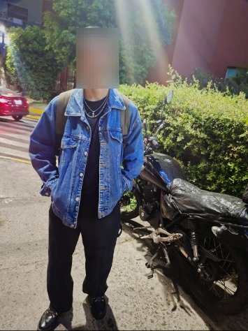 Felipe, in a jean jacket, pauses on sidewalk beside shrub.