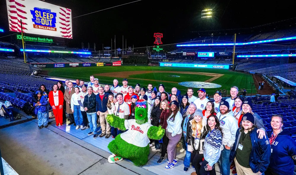 2025 Sleep Out participants at Citizens Bank Park pose with Phillies mascot and with the baseball field and scoreboard in the background.