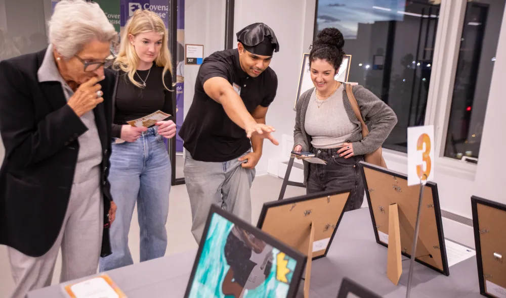 CHNY youth artist in beret explains a piece of art he created to patrons standing around a table with various works.