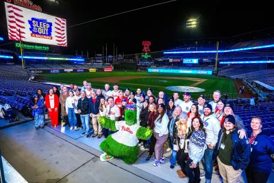 2025 Sleep Out participants at Citizens Bank Park pose with Phillies mascot and with the baseball field and scoreboard in the background.