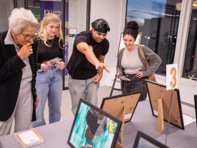 CHNY youth artist in beret explains a piece of art he created to patrons standing around a table with various works.