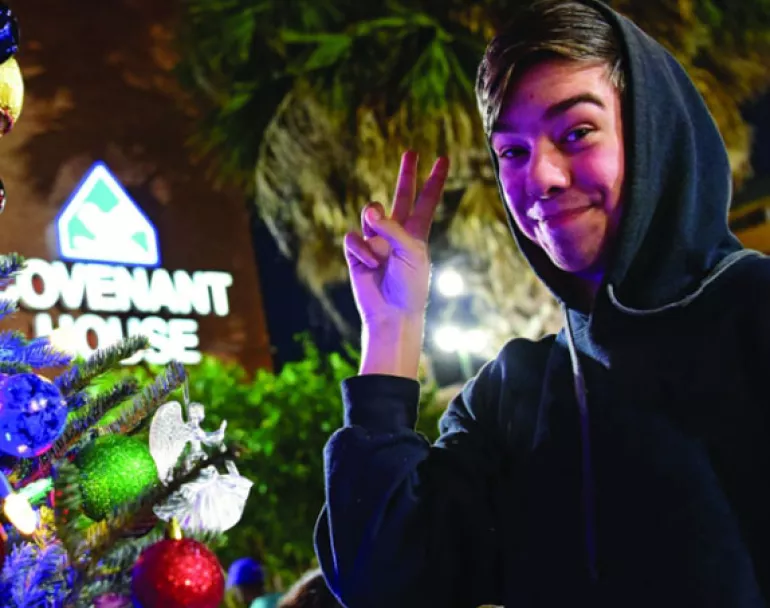 Covenant House former homeless youth making a peace sign next to Christmas Tree | Triple Match Your Donation today!