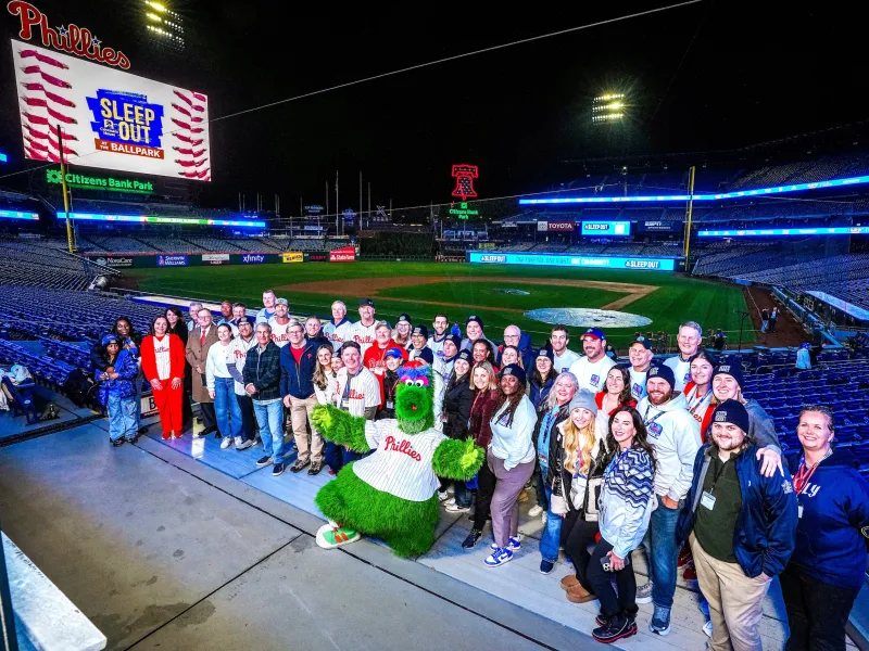2025 Sleep Out participants at Citizens Bank Park pose with Phillies mascot and with the baseball field and scoreboard in the background.